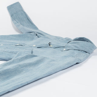 Drake's light blue chambray button-down shirt laid folded on a pale surface, showing the cuff and hem.